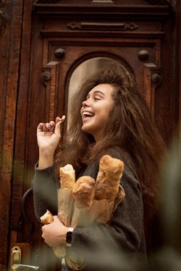 Kız elinde bir baget tutuyordu. Başka tarafa bakıyor ve gülüyor. Kız bir ceket giyiyor. Kalın saçları ve kabarık saçı var. Arkasında kahverengi bir kapı var.