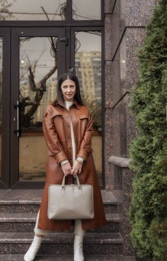 40 yaşın üzerinde zarif bir kadın laptop çantasıyla binadan çıkıyor. O kahverengi bir ceket giyiyor, dizüstü bilgisayar çantası tutuyor. O, beyaz ayakkabılar içinde zarif. O bir ofiste çalışan ince, bakımlı bir kadın..