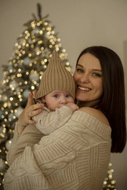 Bebek tutan bir kadın. Kız çocuğunu kollarında tutuyor. Çocuk bej bir el örgüsü şapka giyiyor. Anne ve çocuk mutlu, gülümsüyorlar. Noel 'i kutlarlar. 