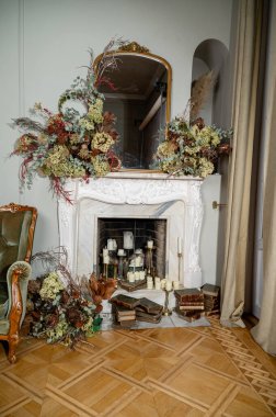 Şömineli oturma odası. Beyaz taştan yapılmış şömine. Yerde ahşap parke var. Şöminenin etrafında bir sürü dekor, yerde mumlar, ayrıca eski kitaplar.