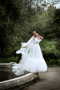 Ağaçların ve eski çeşmelerin arka planında uzun peçeli güzel bir gelin. Uzun beyaz elbiseli esmer kız, pürüzsüz saçları var, arkasında eski bir çeşme var. Duvak yere ulaşır