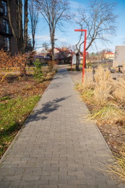 Yürüme yolu, fayanslarla döşenmiş. Yol yerleşim alanından geçiyor. Konut binası ve yeşil çimlerin yanında. Güneşli bir gün.