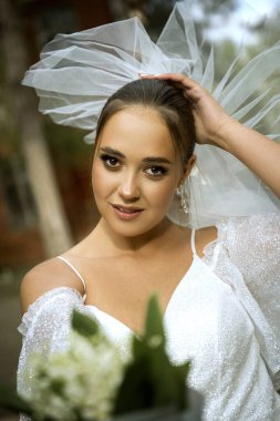 Gelin beyaz bir elbise içinde, omuzları açık, duvağı açık, vadide bir buket zambak. Esmerin topuz şeklinde pürüzsüz bir saç stili var. Kameraya bakıyor.,