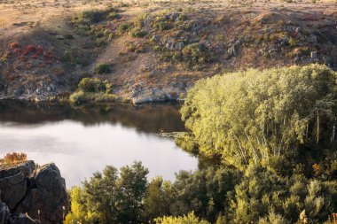Dik kıyılar arasında büyük bir nehir akıyor. Nehrin kıyısındaki sonbahar ormanı. Granit taş kayalıkları. Nehrin sakin su yüzeyinde. Güney böceği, Ukrayna
