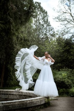 Ağaçların ve eski çeşmelerin arka planında uzun peçeli güzel bir gelin. Uzun beyaz elbiseli esmer kız, pürüzsüz saçları var, arkasında eski bir çeşme var. Bereketli duvak rüzgarda dalgalanıyor