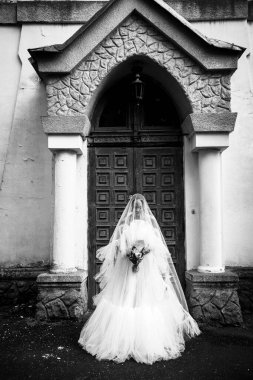 Beyaz elbiseli gelin şifon peçeli ve vadi çiçekli zambak. 18. yüzyıl kilisesinin arka planında uzun elbiseler içinde dik duran kız. siyah beyaz fotoğraf