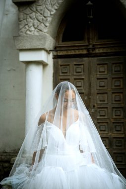 Gelin beyaz bir elbise içinde, omuzları açık, duvağı açık, vadide bir buket zambak. Esmerin topuz şeklinde pürüzsüz bir saç stili var. Gelin uzun bir elbiseyle peçeyle örtülmüş.. 