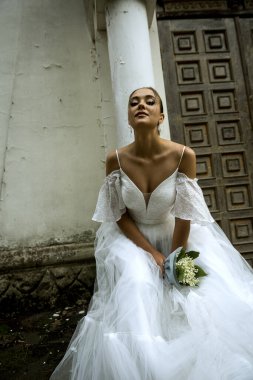 Gelin beyaz bir elbise içinde, omuzları açık, duvağı açık, vadide bir buket zambak. Esmerin topuz şeklinde pürüzsüz bir saç modeli var. Gelin, arkasında 18. yüzyıl kilisesi olduğu için mutlu.