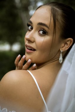 Beyaz peçeli bir gelinin yakın çekimi. Güzel esmer kız düzgün saç stili ve şık makyajlı. Yüzünün yanında da beyaz, zarif bir örtü vardır. Kız gülümser, gelin çerçeveye bakar.