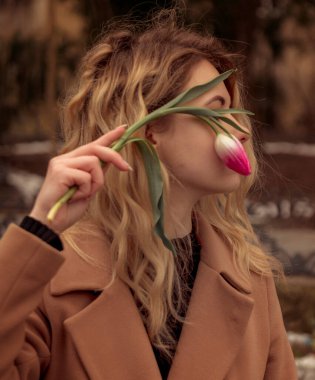 Yüzünde çiçek olan sarışının yakın çekimi. Yüzünü kaplayan laleler. Kızın portresi, yüzünün yanında pembe bir lale tutuyor. Bej bir ceket giymişti. Uzun dalgalı saçları var..