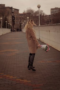 Sarışın ve şehirde bir buket pembe lalesi var. Kız rüzgarda buklelerle dans ediyor. Çerçeveye bakar ve gülümser. O, bej rengi klasik bir ceket giydi.