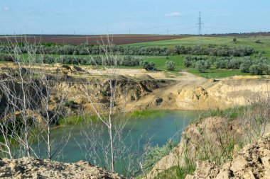 Taş ocağında kocaman mavi bir göl. Etraftaki yaprak döken orman ve tarlalar. Ufuk görünür durumda. Tarlaların arasında yapay bir göl. Ön planda kuru otlar