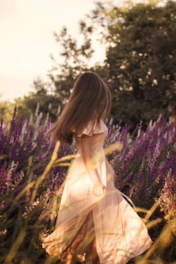 Çiçek açan bir tarlanın ortasında açık renkli, uzun elbiseli ince bir kız. Pembe ve mor çiçekler, lupin ve lavanta, tarlada çiçek açıyor. Kız doğada dans ediyor. Uzun saçları var..
