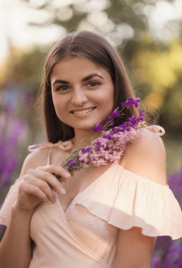 Elinde bir buket yabani çiçekle bir kızın yakın çekimi, gülümsüyor. Kameraya bakıyor. Ellerinde bir buket yabani çiçek var. Elbise yumuşak pembe..