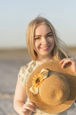 Close-up of a blonde girl with a straw hat in her hands, she laughs and looks at the camera. The hat is decorated with sunflowers. Summer, sunny weather clipart