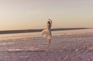 İnce sarışın kız tuz gölünün etrafındaki deniz tuzu yüzeyinde duruyor. Genel plan, batan güneş tarafından aydınlatılması, şifon elbisesinin titremesi. Dans ediyor. Fotoğrafta.