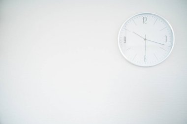 Beyaz bir duvarda beyaz yuvarlak bir saat. Saat sağ üst kısma yerleştirilmiş. Bir yazıt için boşluk. Saat 10.18 gösteriyor.