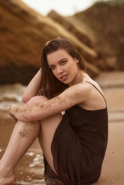 Esmer kız kumlu bir sahilde oturuyor. Kızın uzun saçları var. İnce siyah bir elbise giyiyor. Etrafında büyük sarı kayalar ve deniz vardır. Elbisesi ıslak. Kız çok düşünceli.