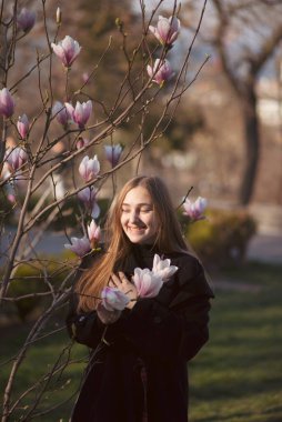 Genç sarışın kız ve çiçek açan pembe manolya. Güneş ışınlarında gülen mutlu kız. Parkta çiçek açan bir ağacın yanında duruyor. Uzun düz saçları ve koyu bir paltosu var..