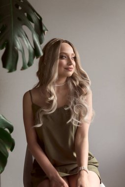 İnce elbiseli zarif sarışın kızın yakın çekimi minimalist iç mekanda sandalyede oturuyor. Kızın uzun açık renk saçları var. Düşünceli bir şekilde başka yere bakıyor. Işık duvarının arkasında, canavarın yanında. yeşil elbise