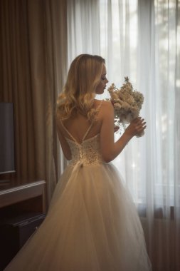 Sırtında bir buket çiçekle, uzun bir gelinlik giymiş bir gelinin silueti. Güzel, genç bir sarışın bir otel penceresinin arkasında duruyor. Beyazdan yapılmış uzun ve şık bir elbise.