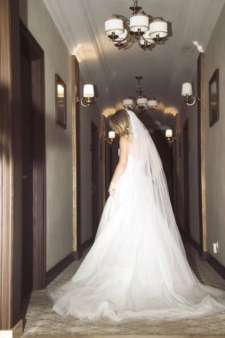 Güzel sarışın gelin otel koridorunda peçeli şık elbiseli, arka manzaralı. Çıplak omuzları ve şık dalgalı saç stili ve makyajlı dantelli gelinlik. Trenli ve uzun peçeli elbise.