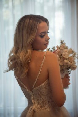 Gelinlerin siluetinin arkası uzun bir gelinlik içinde elinde buketle yakın plan çekilmiş hali. Güzel genç bir sarışın otel penceresinin önünde duruyor.