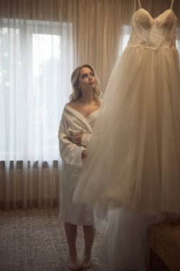 Güzel kız bir otel odasında gelinliğine bakıyor. Beyaz cüppeli ve makyajlı bir kız düğün gününe hazırlanıyor perdenin önünde duran bir insan..