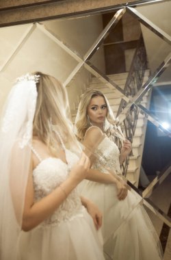 Otelde uzun gelinlik içinde güzel sarışın gelin. Gelin aynaya bakıyor, dalgalı sarı saçları ve açık omuzları var. Elbise ve şifondan yapılmış peçe