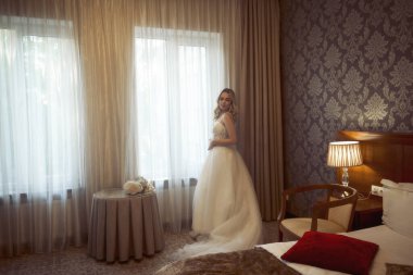 Beyaz elbiseli ve trenli sarışın gelin pencerenin yanında duruyor. Şık bir gelin, şifon ve dantelden yapılmış şık bir elbise içinde gelinin güzel bir saç stili ve makyajı olan düğün günü.