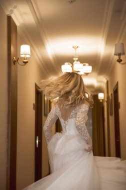 Uzun beyaz elbiseli güzel sarışın gelin otel koridorunda, arka manzaralı. Korseli ve omuzları açık şık bir elbise. Açık renk saçlar ve şık bir stil..