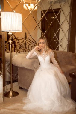 Uzun beyaz elbiseli güzel sarışın gelin, bir otelde oturuyor, bir ayna duvarının arkasında. Uzun kollu, şifon ve dantelden yapılmış şık bir elbise. Açık renk saçlı ve şık.