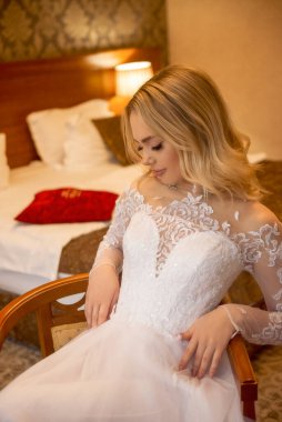 Otel odasında gelinlik giymiş güzel sarışın gelinin yakın çekimi. Uzun kollu, şifon ve dantelden yapılmış şık bir elbise. Dalgalar halinde sarı saçlar. Sandalyede oturuyor.