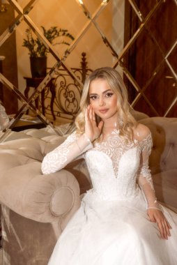 Otelde uzun beyaz elbiseli güzel sarışın gelinin yakın çekimi. Uzun kollu, şifon ve dantelden yapılmış şık bir elbise. Sarı saçlar gevşek ve stil sahibi, büyük küpeler. Kameraya bakıyorum.. 