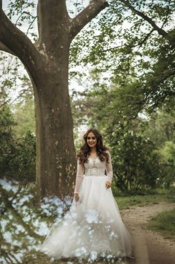 Bir düğün gününde bahar ormanında beyaz elbiseli genç bir gelin. Güzel esmer, uzun ve şık bir planı var. Kocaman ağaçların arasında. Kızın etrafındaki fotoğrafta parla.. 