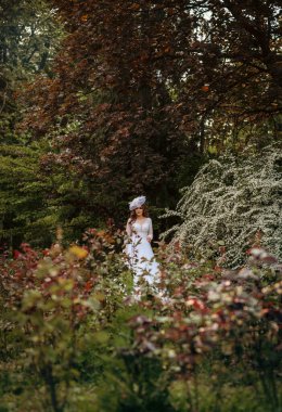 Süzülen Beyaz Elbiseli Zarif Gelin Etrafı Lush Yeşillik ve Serene Garden Ayarlarında Canlı Çiçeklerle çevrili. Beyaz şapkalı, peçeli, klasik tarzda. Çiçek açan ağaçların arasında dalgalı saçları var.