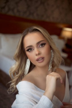 Düğün sabahı bir otelde oturan güzel bir nedimenin portresi. Sarışın, saç stili, uzun saçlı ve makyajlı. Sabahlığını giymiş, düğün günü için hazırlanıyor.. 