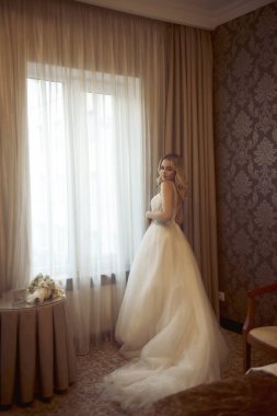Beyaz elbiseli ve trenli sarışın gelin pencerenin yanında duruyor. Şık bir gelinlik içinde, şifon ve dantelden yapılmış. Gelinin düğün günü güzel bir saç stili ve makyajla.
