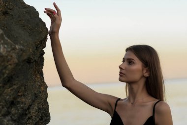 Güzel kız denizin ortasında bir kayanın yanında gün batımının arka planına karşı duruyor. Profilde zarif bir elbise giymiş bir kız.