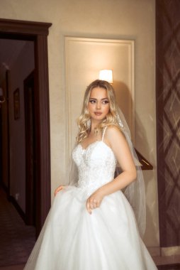 Duvağı olan güzel sarışın gelin şık bir gelinlikle otel lobisinde dikiliyor. Çıplak omuzları ve şık dalgalı saç ve makyajlı, dantelli gelinlik içinde zarif bir gelin..