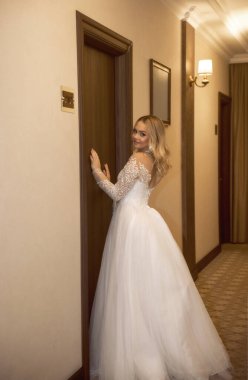 Güzel sarışın gelin otel koridorunda odanın önünde poz veriyor. Uzun gelinlik, dantel ve şifon korsesiyle. Dalgalı sarı saçları, güzel küpeleri var. Gülümsüyor.