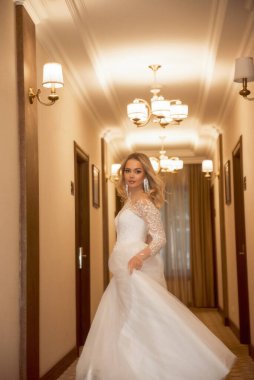 Otel koridorunda poz veren uzun beyaz elbiseli güzel sarışın gelin. Korseli ve omuzları açık şık bir elbise. Sarı saçlar bol ve şık bir stili var. Gülümsüyor ve dans ediyor.