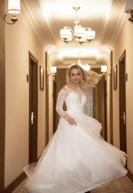 Otel koridorunda poz veren uzun beyaz elbiseli güzel sarışın gelin. Korseli ve omuzları açık şık bir elbise. Sarı saçlar bol ve şık bir stili var. Gülümsüyor ve dönüyor.