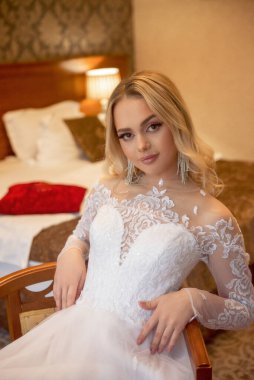 Otel odasında gelinlik giymiş güzel sarışın gelinin yakın çekimi. Uzun kollu, şifon ve dantelden yapılmış şık bir elbise. Dalgalar halinde açık renk saçlar, pahalı küpeler.