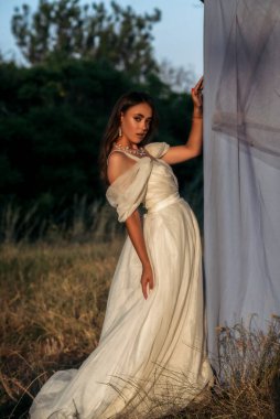 Parlak makyajlı ve uzun beyaz elbiseli güzel bir kadın. Doğada, kumaş arka planının yanında tam gelişim içinde poz veriyor. Dekoratif kozmetik ürünleri, şık kıyafetler.