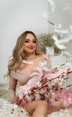 Tüylerin arasındaki güzel sarışın kız. Beyaz tüylerin arasında oturuyor. Etrafından uçuyorlar. Elinde pembe büyük tüyler ve pamuktan dallar tutuyor.