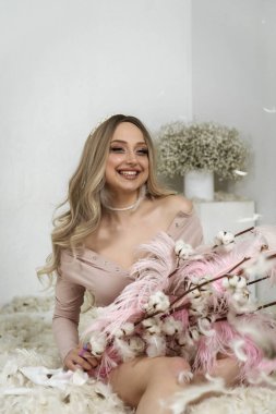 Tüylerin arasındaki güzel sarışın kız. Beyaz tüylerin arasında oturuyor ve gülümsüyor. Tüyler havada uçuşuyor. Elinde pembe büyük tüyler ve pamuktan dallar tutuyor.