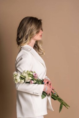 Beyaz takım elbiseli güzel bir kızın stüdyo fotoğrafı. Kız bej bir arka planda duruyor. Elinde çiçekler var. profil resmi