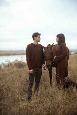 Genç ve sevecen bir adam ve kadın atın yanında at binerken dinleniyorlar. Bir sonbahar tarlasının arka planında şık bir şekilde kahverengi kıyafetler giyiyorlar. Bir atın yanında duruyorlar ve...