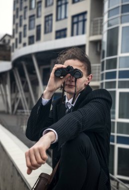 Genç bir adamın portresi modern mimarinin arka planına karşı dürbünle bakıyor. Uzun saçları var, takım elbise giyiyor. Ön manzara, yakın plan. Rahat, huzurlu, düşünceli.